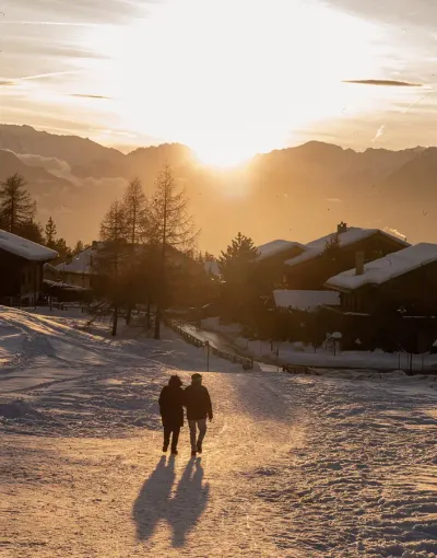 Twee mensen lopen bij zonsondergang over een besneeuwd pad naar een paar chalets
